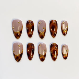 Set of ten almond-shaped false nails. Each nail features a gradient design with a nude base, transitioning to a dark brown tip with gold glitter accents. Small gold studs are evenly spaced across each nail.