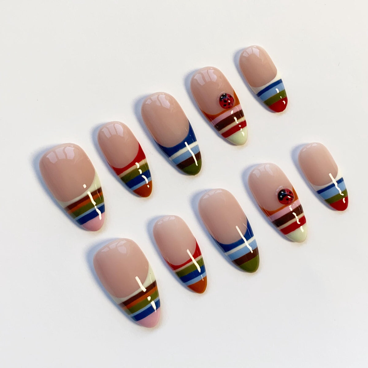 A set of ten artificial nails with a nude base and colorful striped designs. Some nails feature a small ladybug accent. The stripes include red, blue, green, and yellow, creating a vibrant and playful look.