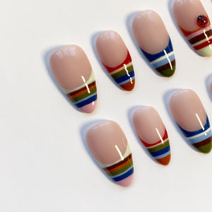 A collection of oval-shaped false nails with a nude base and colorful striped designs. The stripes include red, orange, green, blue, and white. One nail features a ladybug design. The nails are arranged on a white surface.
