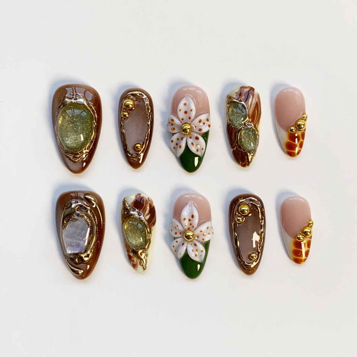 A set of ten artificial nails with various designs. Some nails feature brown and gold accents, while others showcase white floral designs with green bases. Several nails have glittery embellishments and gold studs.