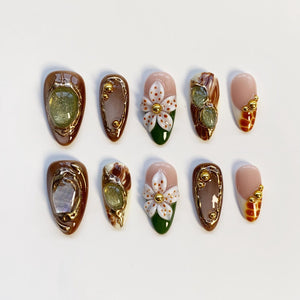 A set of ten artificial nails with various designs. Some nails feature brown and gold accents, while others showcase white floral designs with green bases. Several nails have glittery embellishments and gold studs.