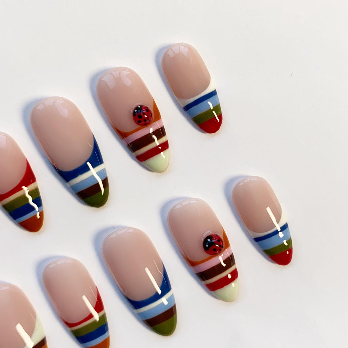Oval-shaped artificial nails with a nude base and colorful striped tips. Some nails feature a small ladybug design. The stripes include red, blue, green, and brown. The nails are arranged on a white surface.