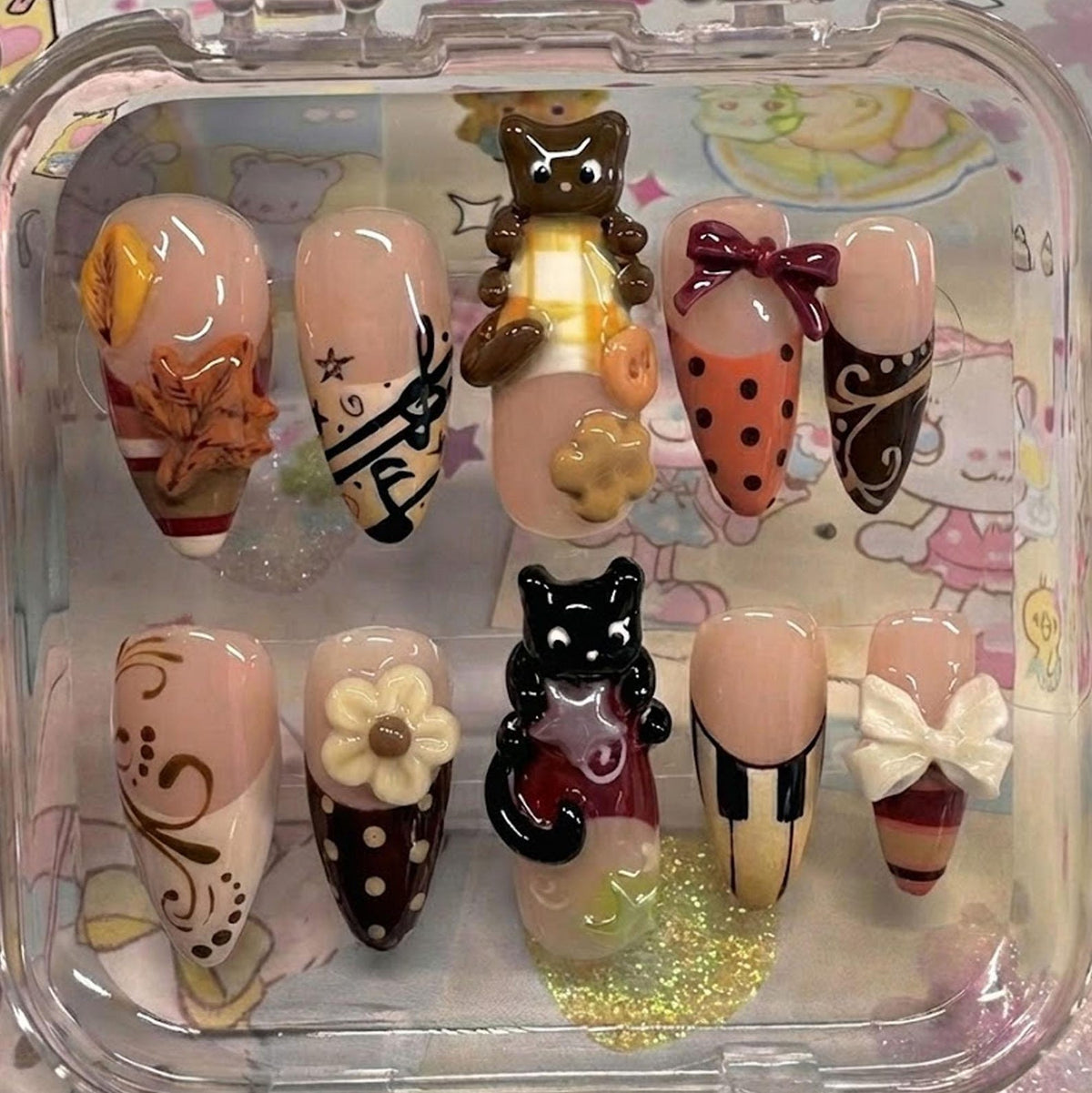 A set of ten artificial nails with various designs. The nails feature a range of colors, including nude, brown, orange, and white, and are adorned with decorative elements such as bows, flowers, and animal figurines. The nails are displayed in a clea