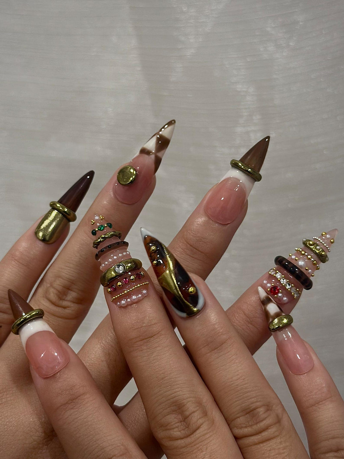 A set of long, pointed artificial nails. The nails feature a variety of designs, including brown and white checkered patterns, gold accents, and decorative rings with pearls and gems. The nails have a natural pink base with white tips.
