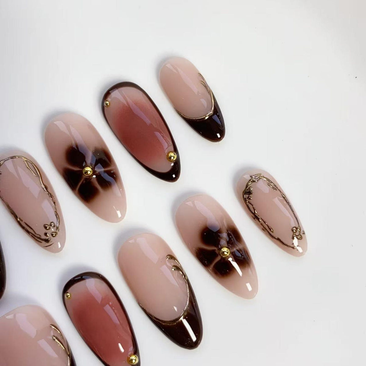 A set of almond-shaped artificial nails. The nails feature a gradient design in shades of brown and nude, with gold accents. Some nails have a floral design, while others have gold lines and small gold studs.