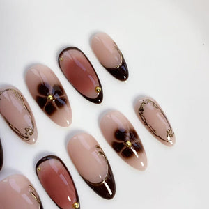 A set of almond-shaped artificial nails. The nails feature a gradient design in shades of brown and nude, with gold accents. Some nails have a floral design, while others have gold lines and small gold studs.