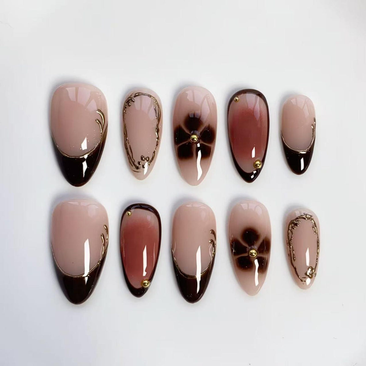 A set of ten almond-shaped false nails. The nails feature a gradient design with a nude base, dark brown tips, and gold accents. Some nails have gold floral designs or small gold studs.