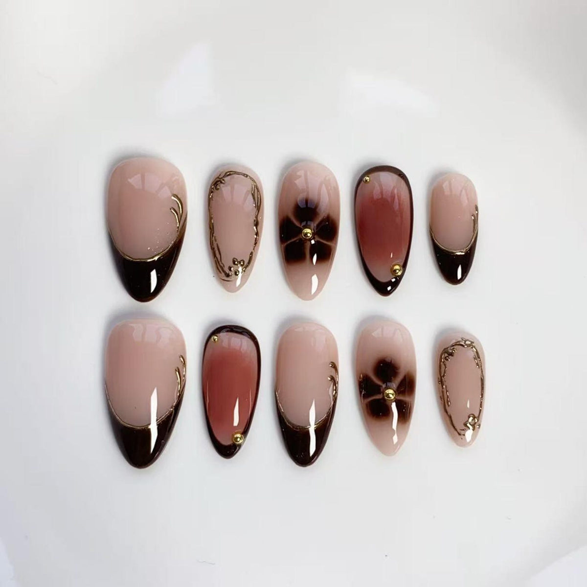 Set of ten almond-shaped false nails. The nails feature a nude base with various designs, including dark brown tips, floral accents, and gold embellishments. Some nails have a dark brown gradient, while others have gold swirls.