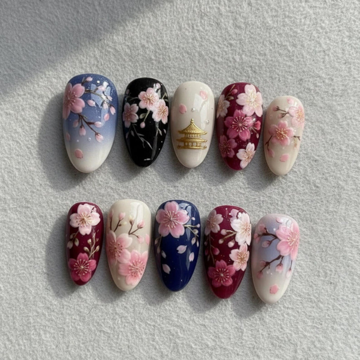 A set of ten artificial nails with a floral design. The nails feature a variety of colors, including blue, black, white, and burgundy, with pink cherry blossom accents. Some nails have a gold pagoda design.