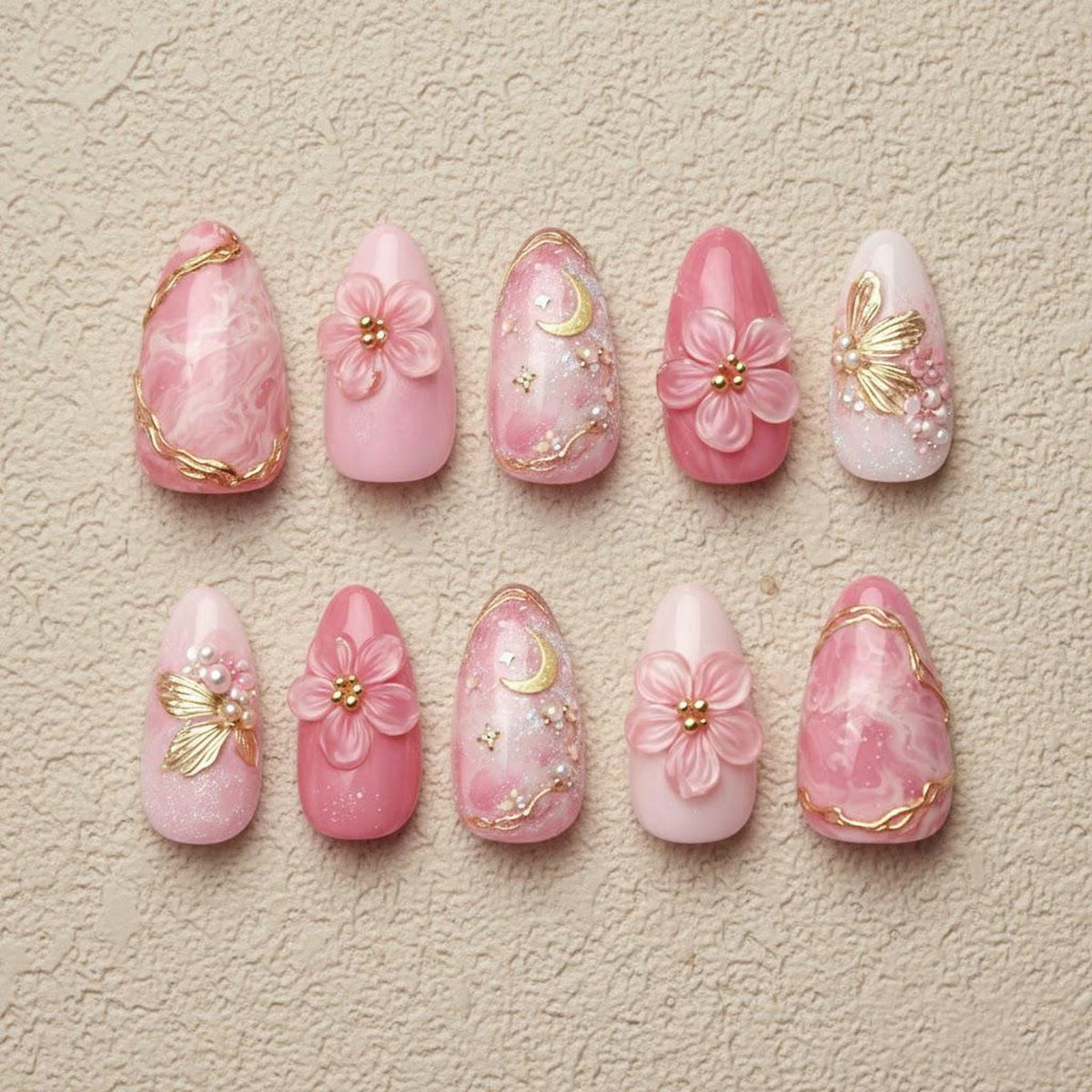 A set of ten artificial nails in various shades of pink. Designs include marble effects, gold accents, floral embellishments, and moon and star details. The nails are oval-shaped and arranged on a neutral surface.