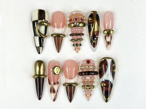 A collection of ten artificial nails in various designs. Some nails feature a checkerboard pattern, while others have a nude base with gold or brown accents. Several nails are adorned with rings and jewels.