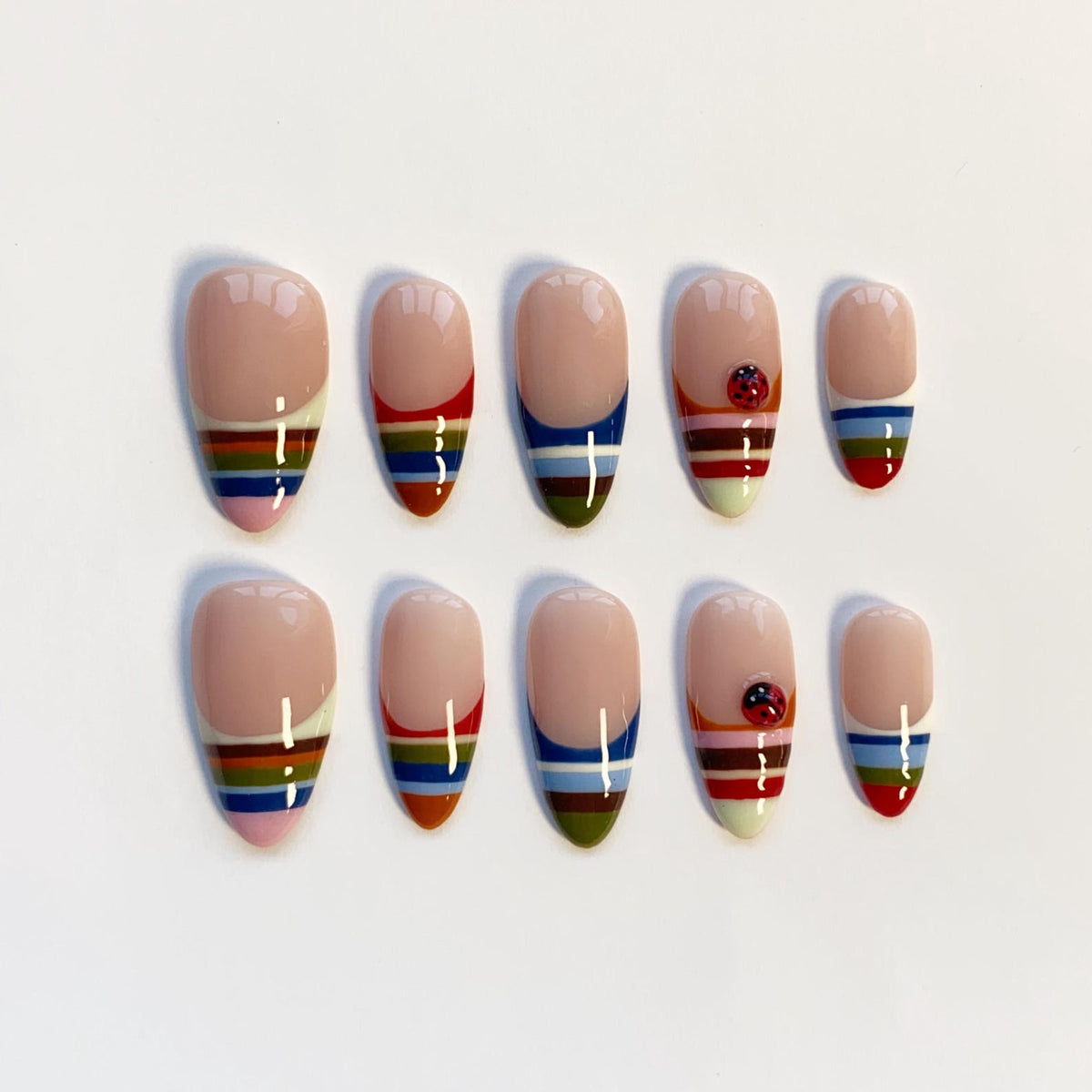 A set of ten almond-shaped false nails. Each nail features a nude base with a colorful striped design at the tip. Some nails have a small ladybug accent. The stripes include red, blue, green, yellow, and pink.