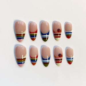 A set of ten almond-shaped false nails. Each nail features a nude base with a colorful striped design at the tip. Some nails have a small ladybug accent. The stripes include red, blue, green, yellow, and pink.