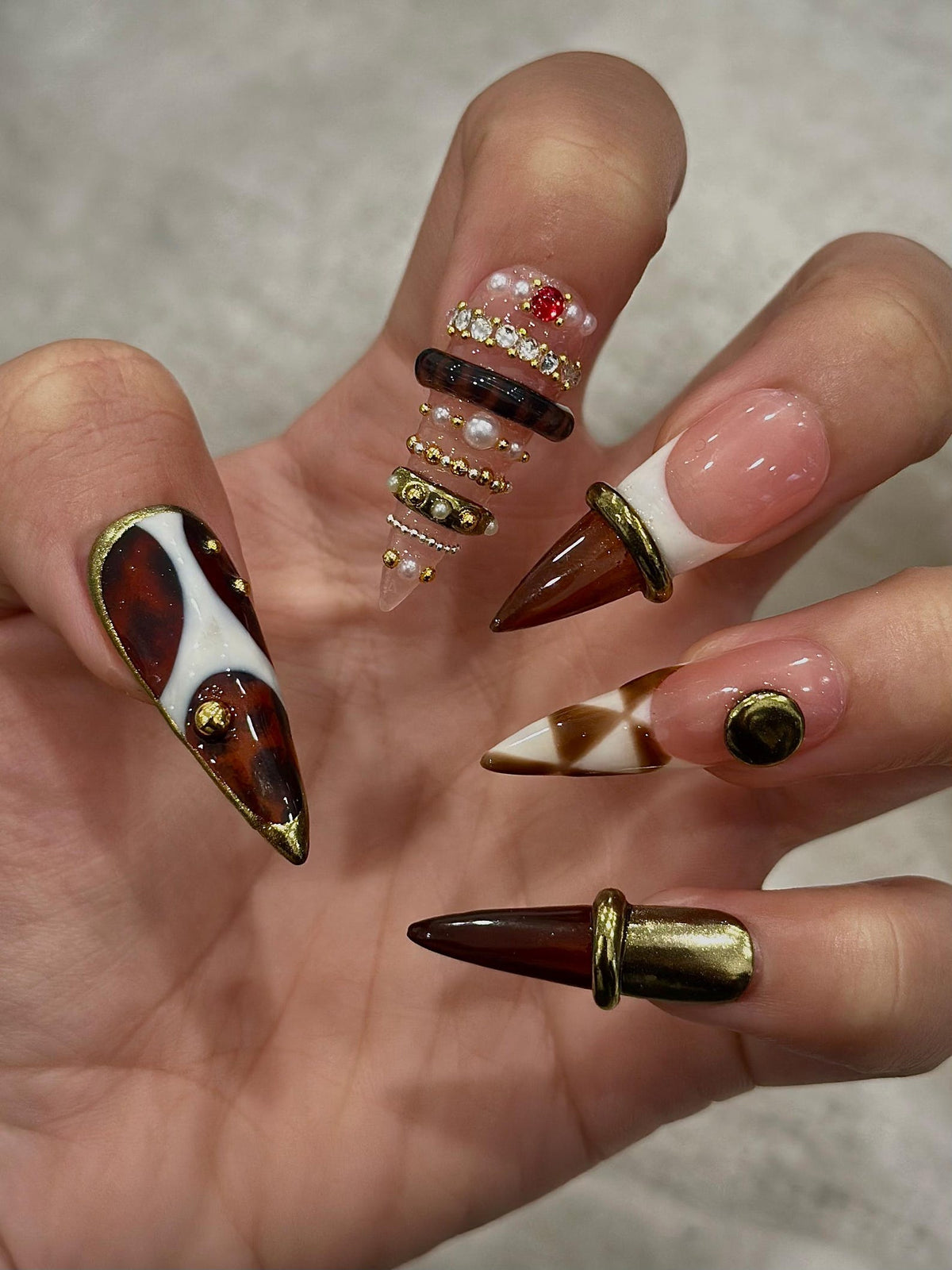 A hand displaying a set of long, pointed acrylic nails. The nails feature various designs, including brown and white patterns, gold accents, and embedded gems. Some nails have a French tip design with a brown cone shape.