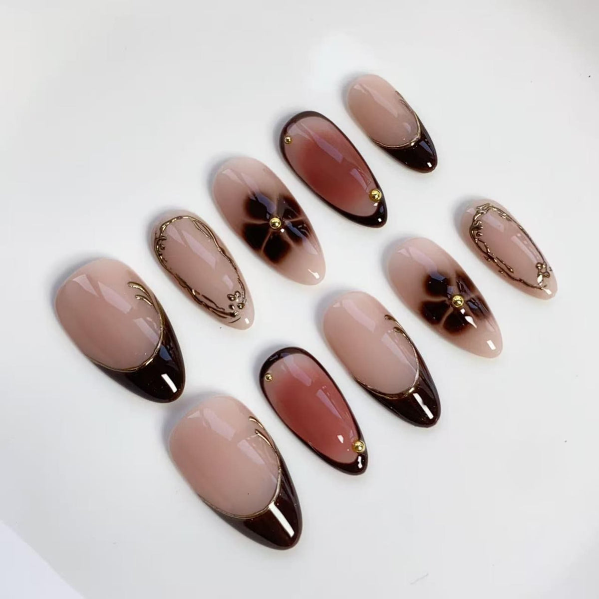 A set of ten almond-shaped artificial nails. The nails feature a nude base with various designs, including dark brown tips, floral patterns, and gold accents. Some nails have a dark brown border, while others have gold embellishments.