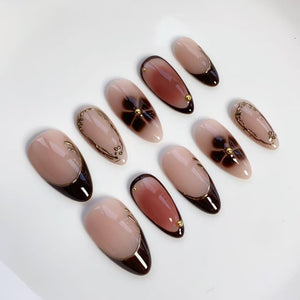 A set of ten almond-shaped artificial nails. The nails feature a nude base with various designs, including dark brown tips, floral patterns, and gold accents. Some nails have a dark brown border, while others have gold embellishments.