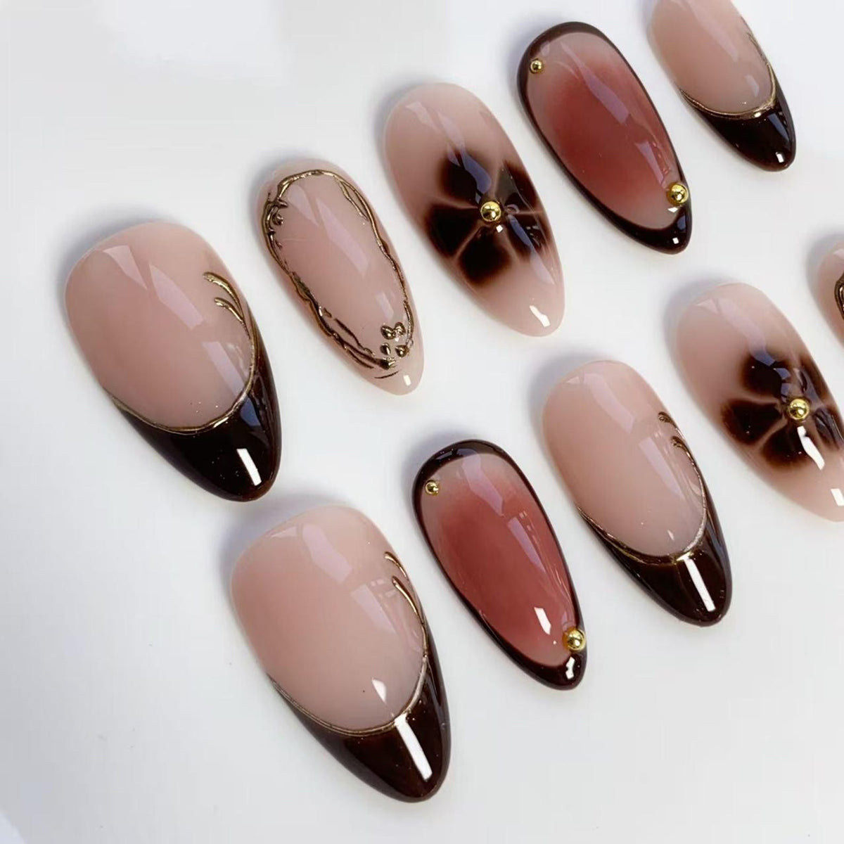 A collection of almond-shaped artificial nails. The nails feature a variety of designs, including a dark brown French tip with gold trim, floral patterns, and gold embellishments. The base color is a nude pink.