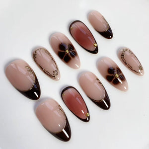 Oval-shaped artificial nails in various designs. Some nails feature a dark brown French tip, while others have a floral design or gold embellishments. The nails are in shades of nude, brown, and gold.
