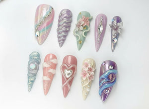 An assortment of ten long, almond-shaped artificial nails. Each nail features unique designs, including swirls, gems, flowers, and textured patterns in pastel colors like pink, purple, and blue. The nails are arranged on a white surface.