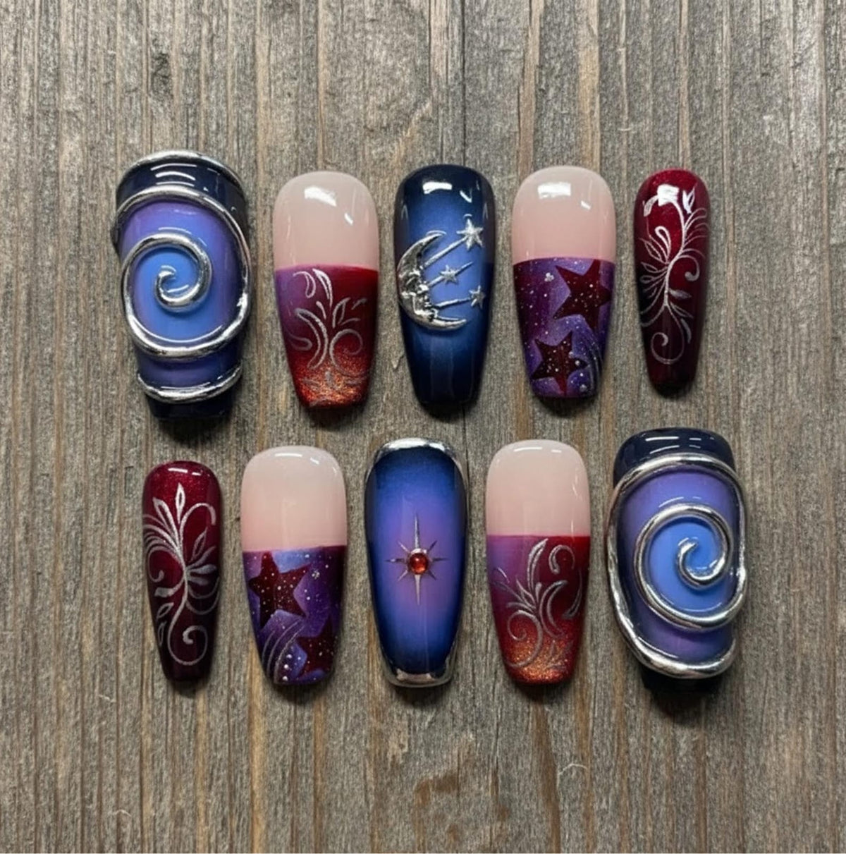 A set of ten artificial nails with various designs. Some nails feature a gradient of colors, including pink, purple, and red. Other nails have silver embellishments, stars, and a moon design. Two nails have a silver swirl design.