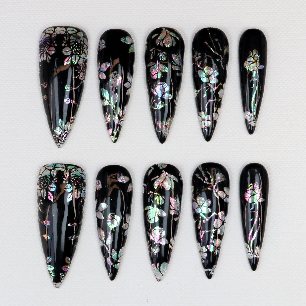 A set of ten black, almond-shaped artificial nails. Each nail features a floral design with iridescent accents. The flowers and leaves have a shimmering, multi-colored effect, creating a striking contrast against the dark background.