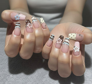 A set of long, square-shaped acrylic nails with a variety of designs. Some nails are light pink with white star accents, while others feature brown, white, and light blue stripes or polka dots. Silver rhinestones add sparkle.