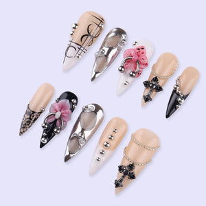 A collection of long, pointed artificial nails in various designs. Some nails feature silver embellishments, floral accents, and cross pendants. Colors include nude, black, white, and silver, with intricate patterns and textures.