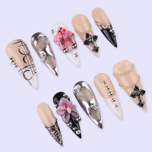 A collection of ten long, almond-shaped artificial nails. Designs include silver metal accents, crosses, floral embellishments, and various patterns. Colors include black, white, nude, and silver, with rhinestones and chain details.