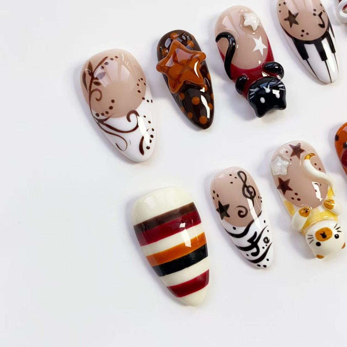 A collection of decorated artificial nails in various designs. Some nails feature brown star accents, musical notes, and cat figures. Others have striped patterns in red, orange, and black. The nails are set against a white background.