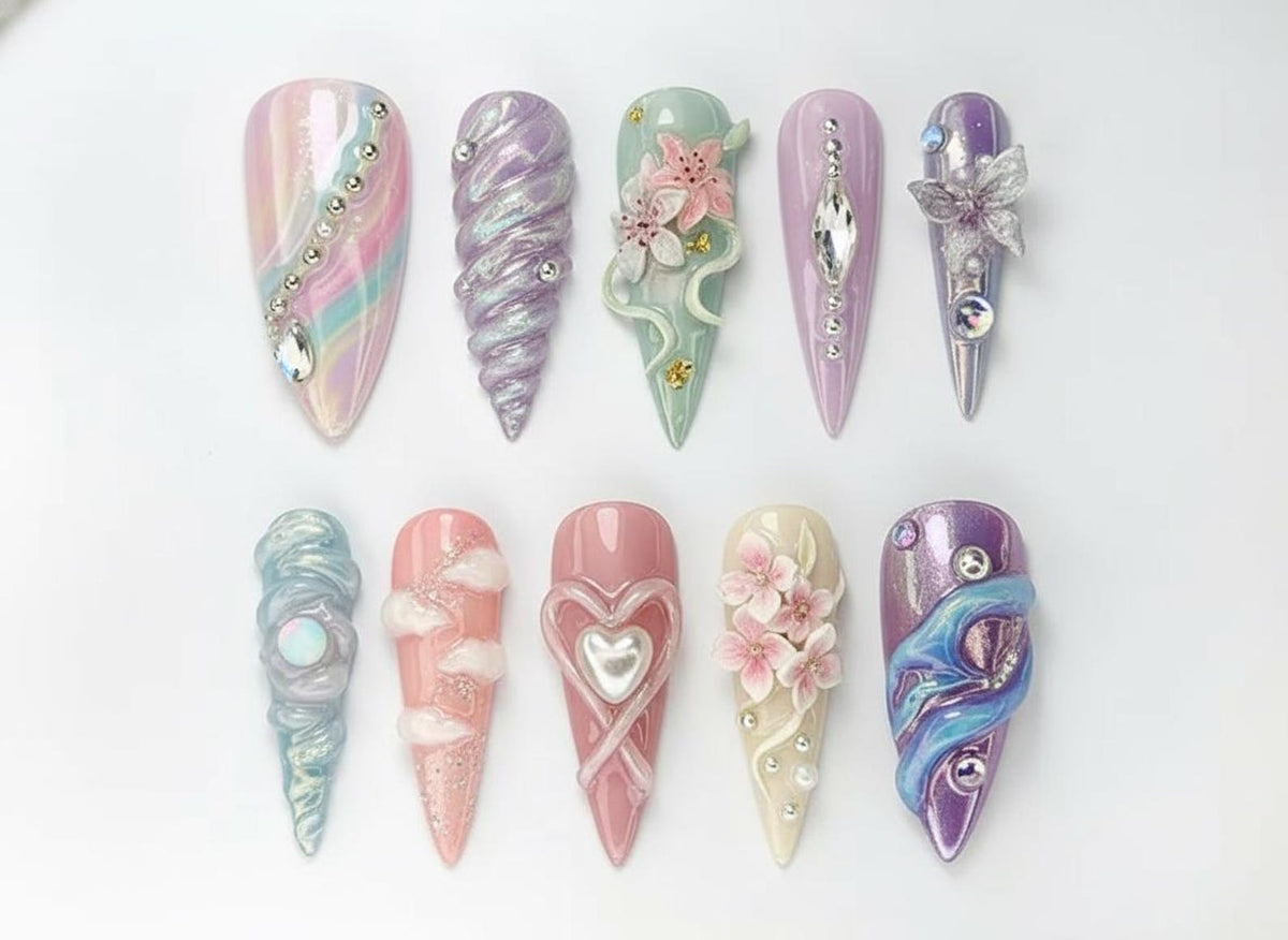 An assortment of ten long, almond-shaped artificial nails. Each nail features unique designs, including iridescent finishes, sculpted elements, and embellishments like rhinestones, pearls, and floral accents. Colors range from pastel pinks and purple
