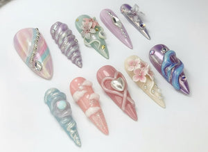 A collection of ten long, almond-shaped artificial nails. Each nail features a unique design, including iridescent finishes, sculpted embellishments, and decorative accents like gems, pearls, and floral details. The color palette includes pastels, pi