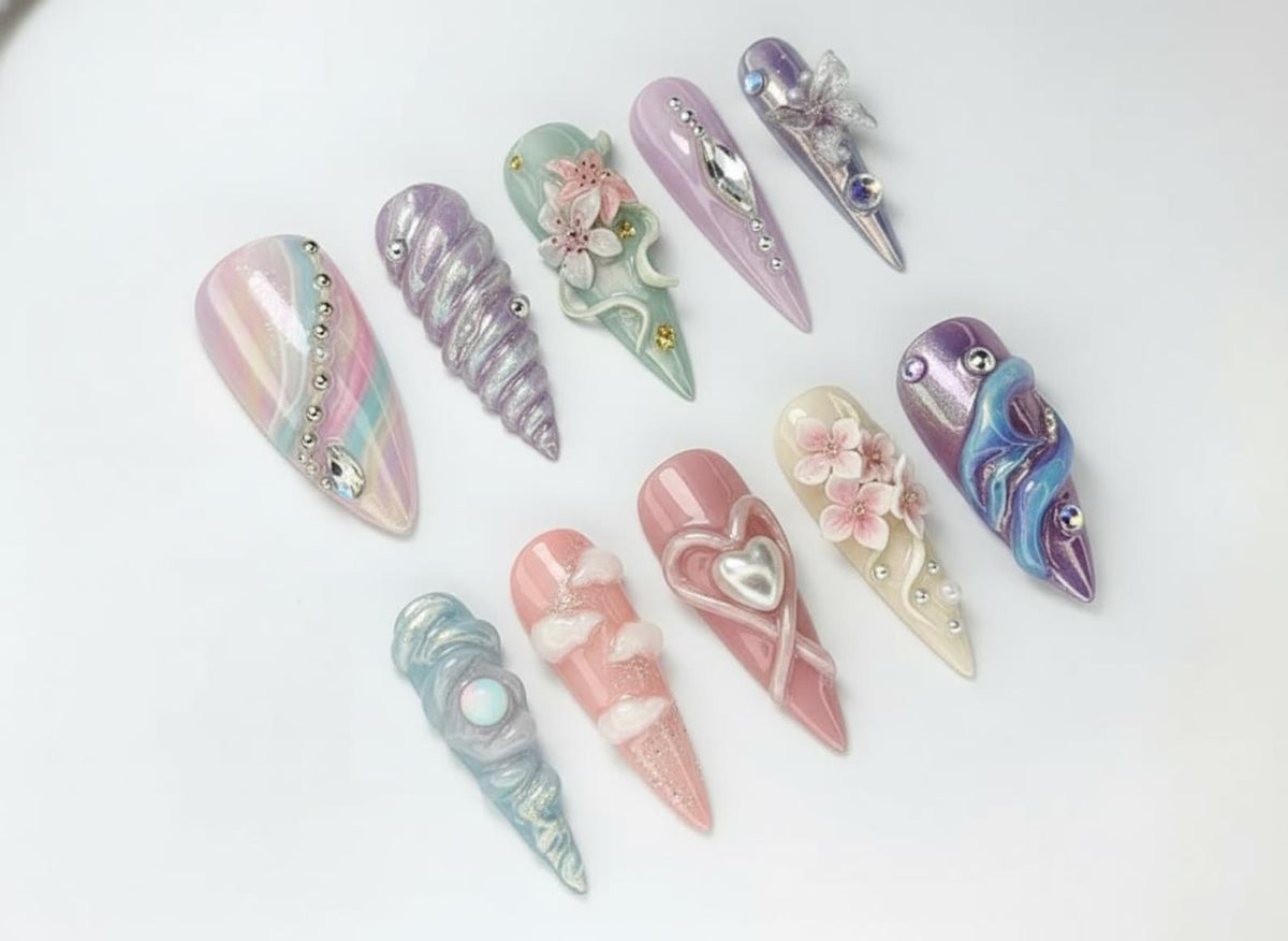 A collection of ten long, almond-shaped artificial nails. Each nail features unique designs, including pastel colors, pearls, floral accents, and 3D embellishments. The nails are arranged on a white surface.