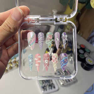 A set of ten artificial nails in a clear plastic case. The nails feature various designs, including swirls, floral accents, and gem embellishments. Colors include pastel pinks, blues, purples, and white.