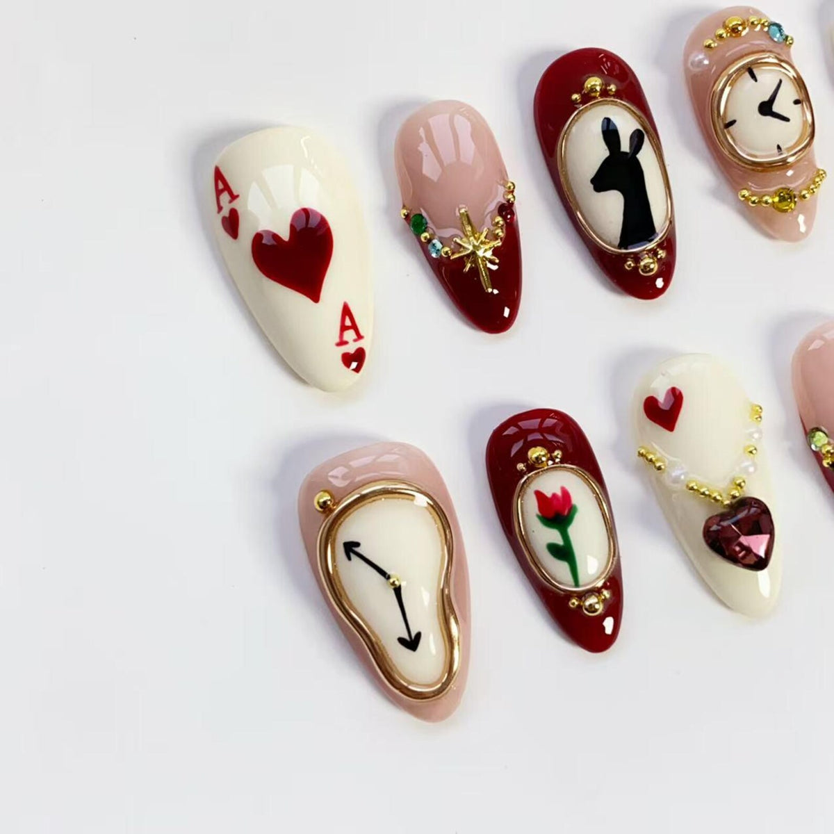 A collection of artificial nails with various designs. One nail features the Ace of Hearts playing card, while others showcase clocks, a deer silhouette, and a rose. The nails are painted in shades of red, white, and pink, with gold accents.