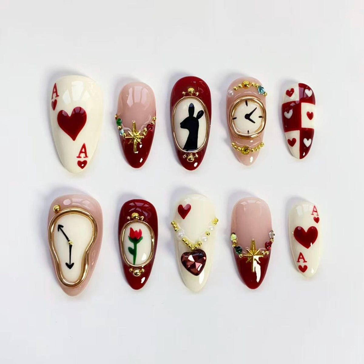 A set of ten almond-shaped artificial nails with various designs. The designs include playing card suits, clocks, a silhouette of a rabbit, and floral and jewel accents. The nails are painted in shades of red, white, and pink.
