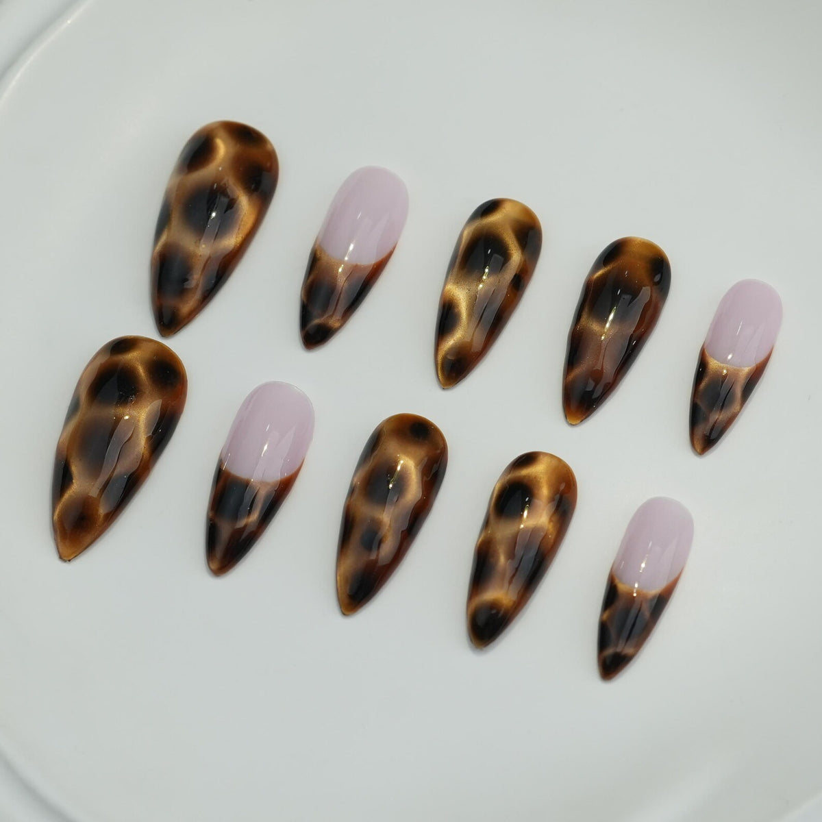 A set of ten artificial nails. The nails feature a glossy, almond shape. Some nails have a brown and tan tortoiseshell pattern, while others have a light pink tip with a brown base. The nails are arranged on a white surface.