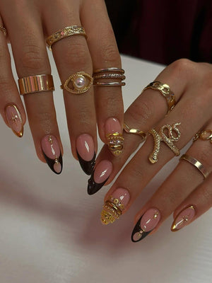 A collection of gold rings in various designs, including a snake, eye with a pearl, and bands, adorn fingers with almond-shaped nails. The nails feature a pink base with black tips and gold accents, showcasing a stylish manicure.