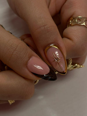 Close-up of manicured nails with a gold border. One nail features a starburst design with a small gem. The other nails have a pink base with a dark brown tip. Gold rings adorn the fingers.