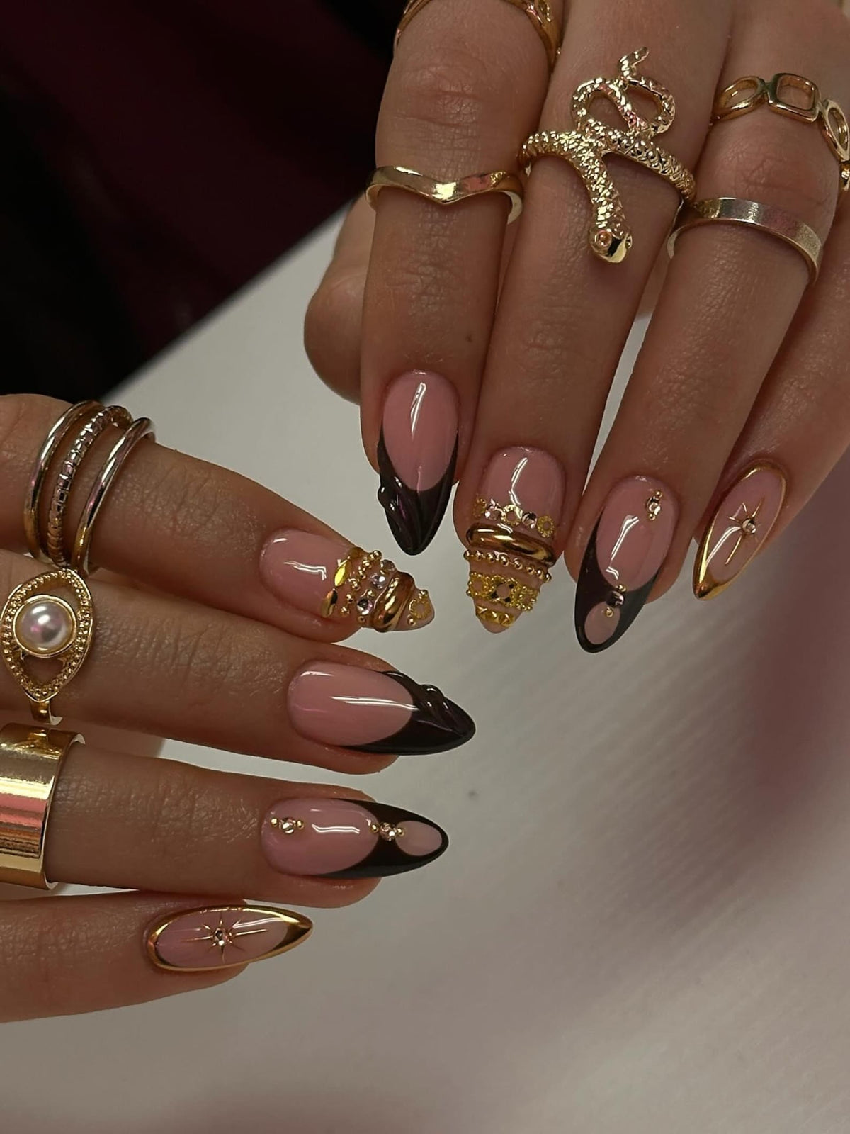 Close-up of manicured nails with a pink base and dark brown tips. Some nails feature gold embellishments and a gold star design. The image also shows various gold rings.