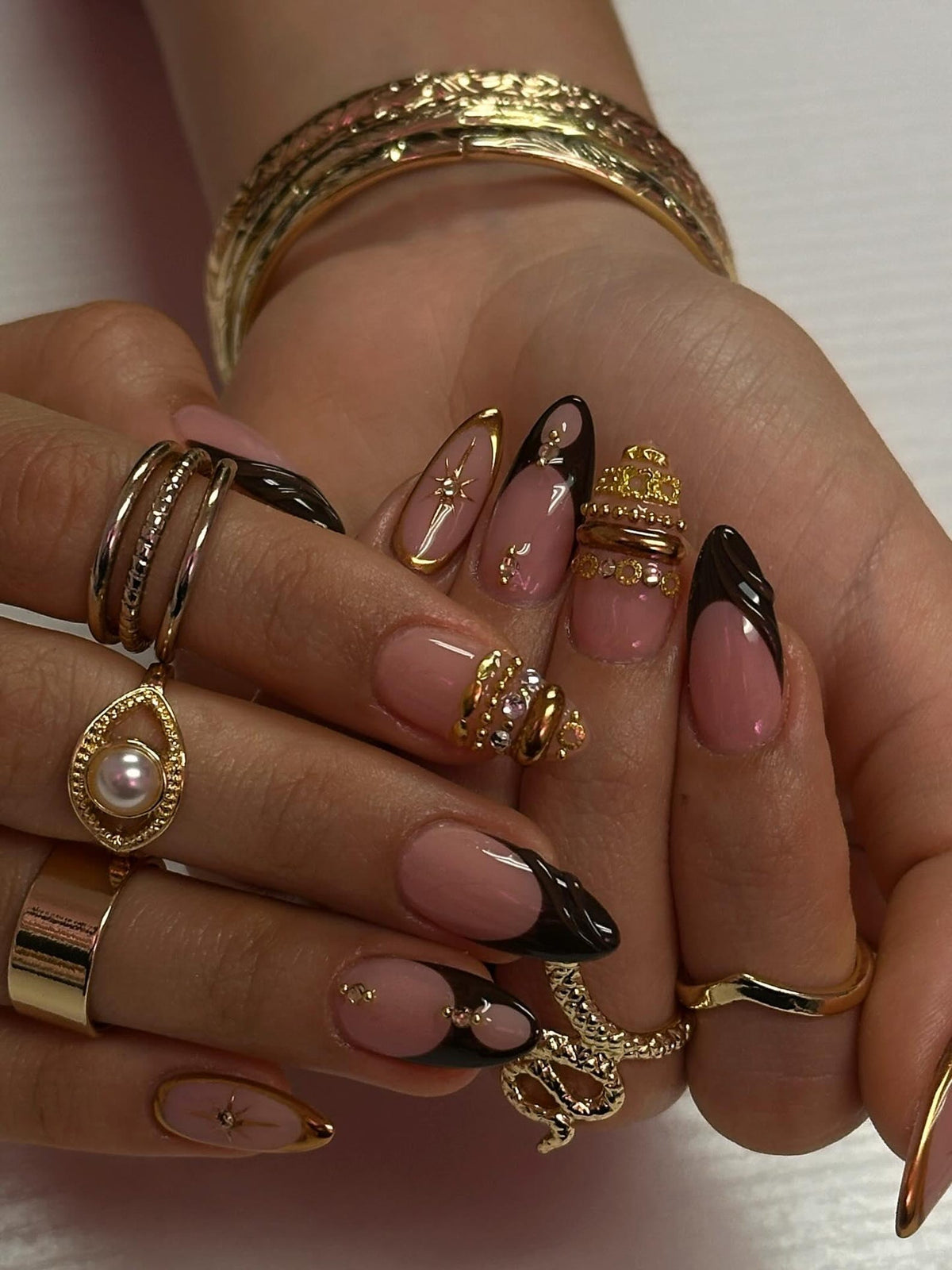A hand adorned with gold rings and a gold bracelet. The nails are painted with a French manicure design, featuring a nude base with black tips and gold accents. Some nails have gold embellishments.