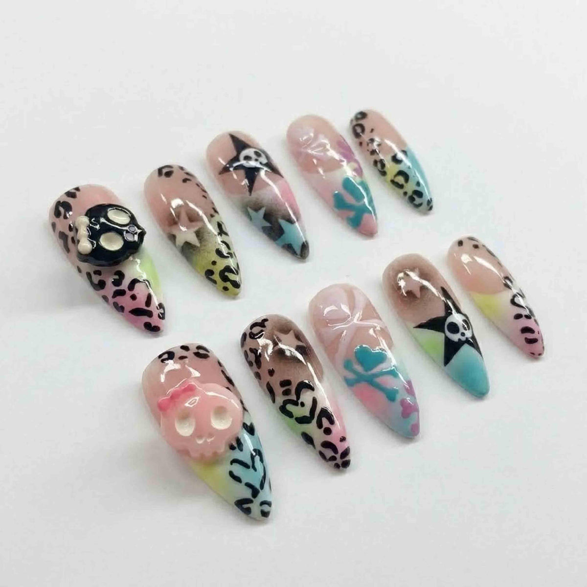 A set of ten long, almond-shaped artificial nails. The nails feature various designs, including leopard print, skulls, stars, and pastel color gradients. Some nails have 3D skull embellishments, and others have painted crossbones.