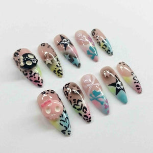 A set of ten long, almond-shaped artificial nails. The nails feature various designs, including leopard print, skulls, stars, and pastel color gradients. Some nails have 3D skull embellishments, and others have painted crossbones.