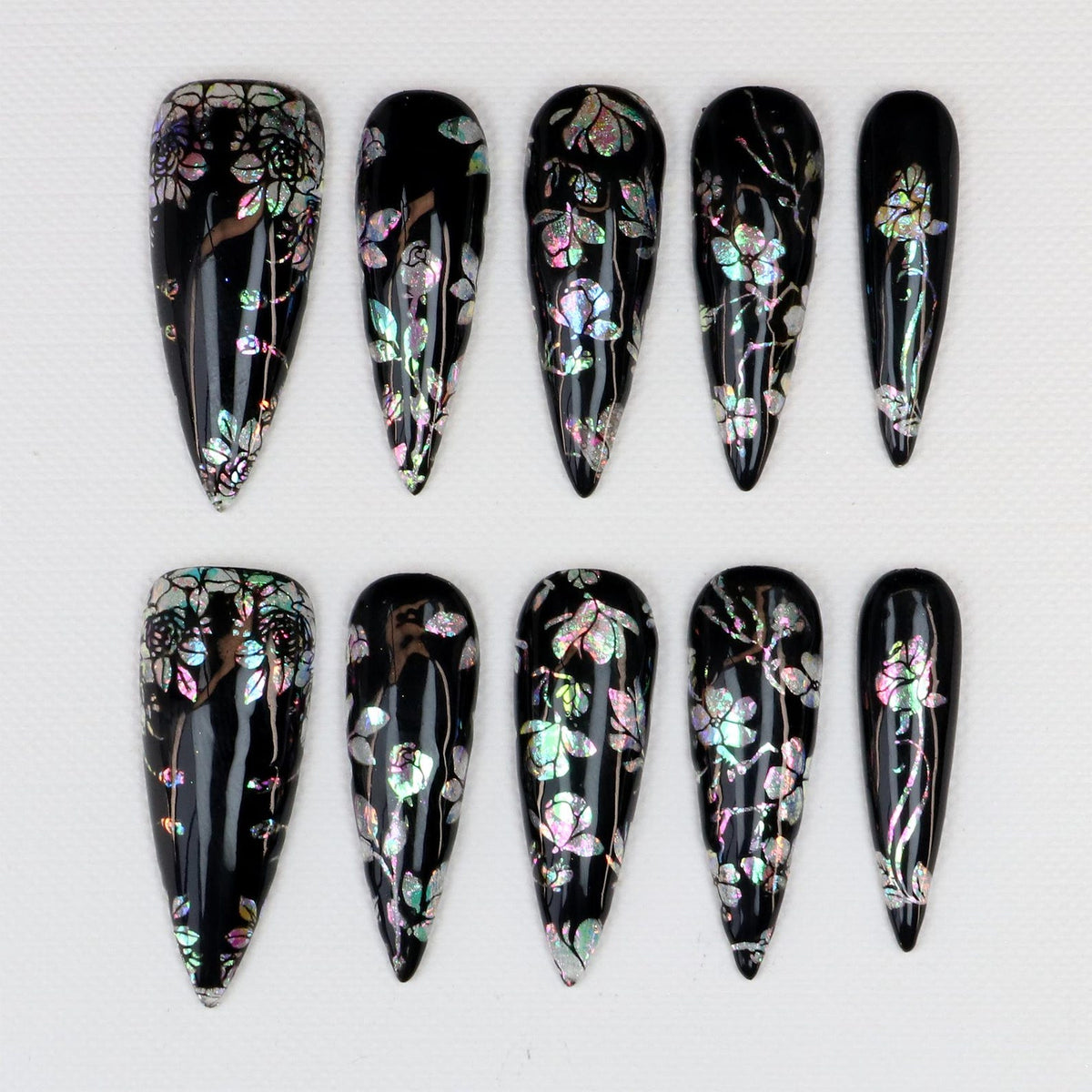 Set of ten black press-on nails with iridescent floral designs. Each nail has a glossy finish and a different floral pattern in shades of pink, green, and silver. The nails are almond-shaped.
