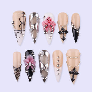 A collection of ten artificial nails in various designs. Some nails are nude-colored with silver studs, black accents, or lace patterns. Others feature silver metal accents, pink floral designs, or black cross pendants. The nails are long and pointed
