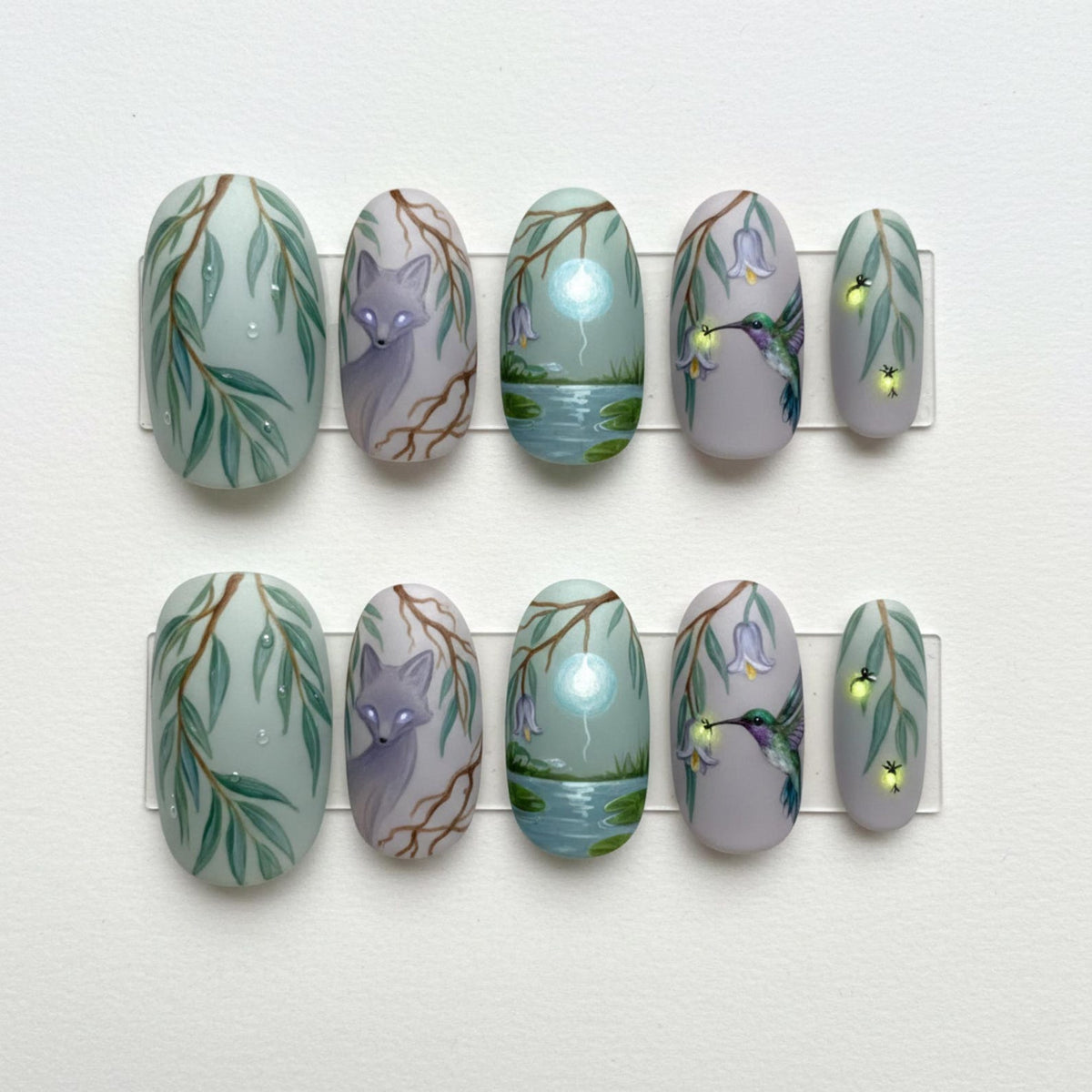 Set of ten oval-shaped false nails with nature-themed art. Designs include willow branches, a fox, a pond scene with a glowing orb, a hummingbird, and fireflies. The nails are painted in shades of green, gray, and purple.
