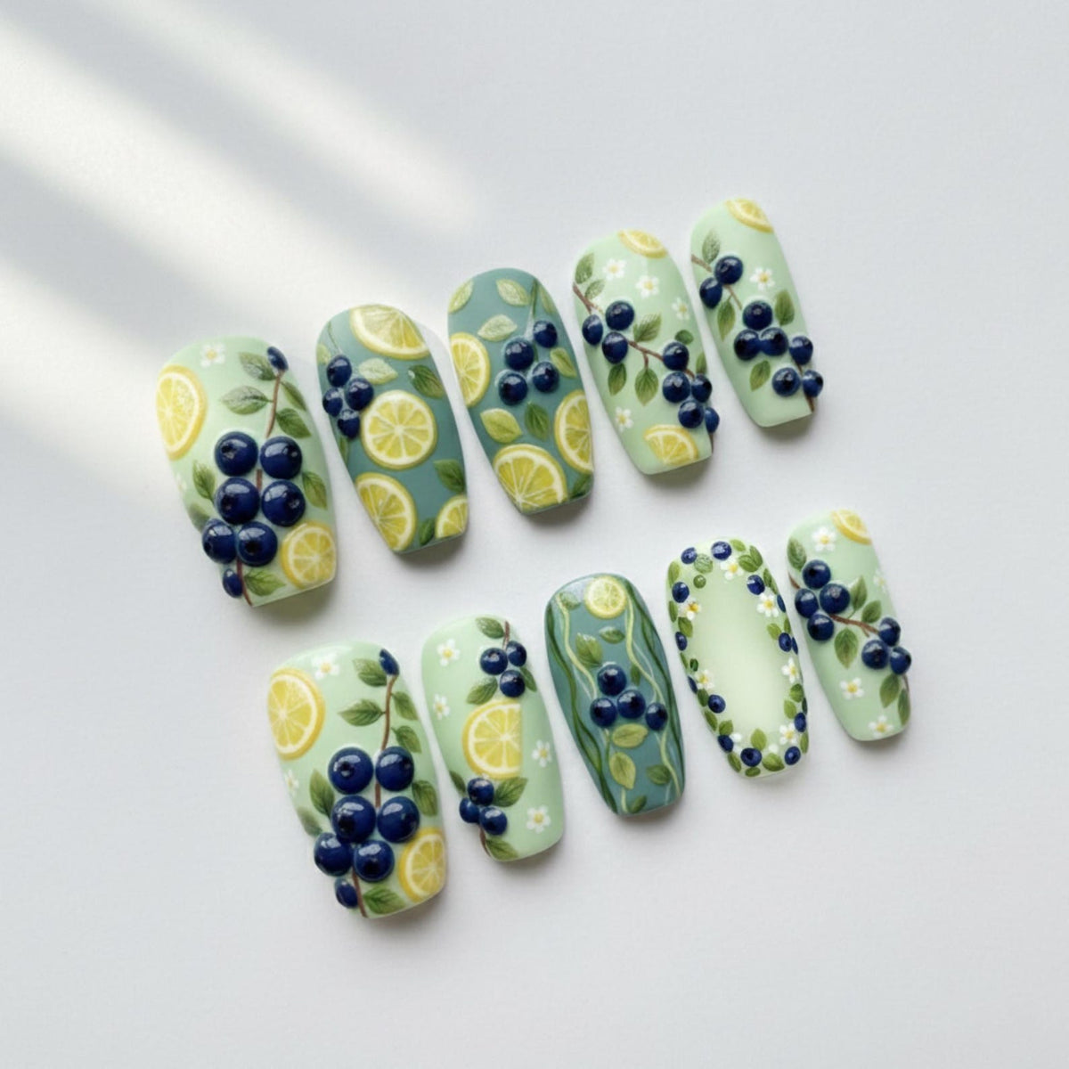A set of ten artificial nails with a light green base, decorated with lemon slices, dark blue berries, and green leaves. Some nails feature a solid color, while others have a patterned design.