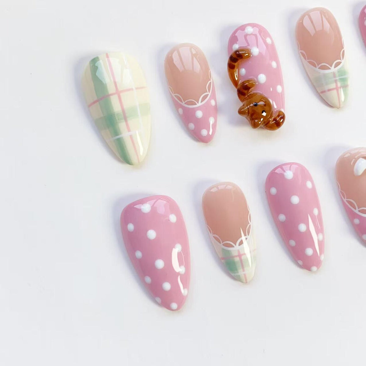A collection of almond-shaped artificial nails. Designs include pink with white polka dots, plaid, and a 3D brown bear. Some nails have a nude base with white lace detailing. The nails are arranged on a white surface.