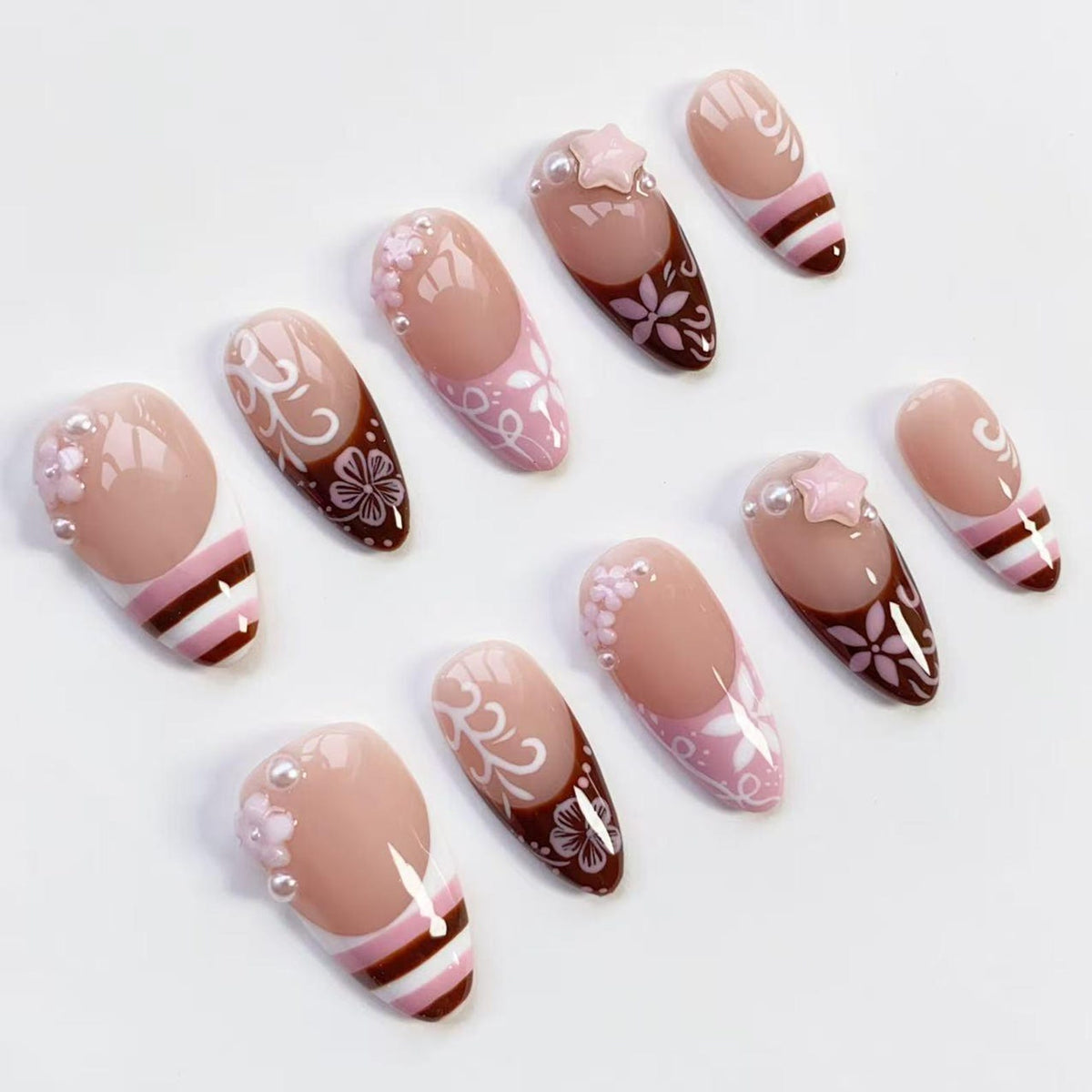 A collection of artificial nails in various designs. The nails feature a nude base with pink, white, and brown accents. Some nails have floral patterns, pearl embellishments, and star-shaped decorations.