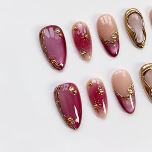 Oval-shaped artificial nails in various designs. Some nails are a deep burgundy with gold trim and gold embellishments. Others feature a gradient from light pink to burgundy with gold starburst and dot accents.