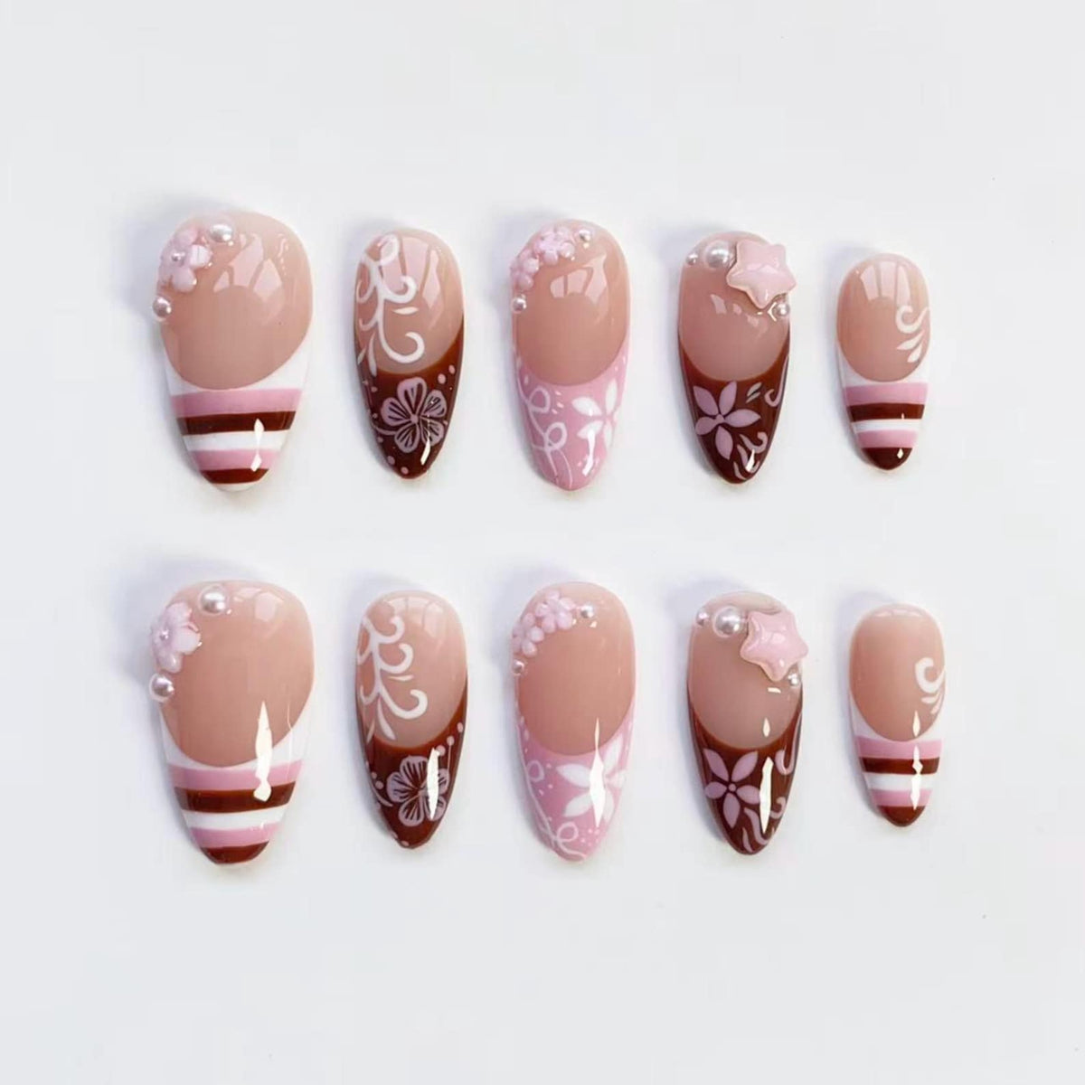 A set of ten artificial nails in various designs. The nails feature a nude base with accents of pink, white, and brown. Some nails have floral patterns, pearls, or star-shaped embellishments. Others have striped or swirl designs.